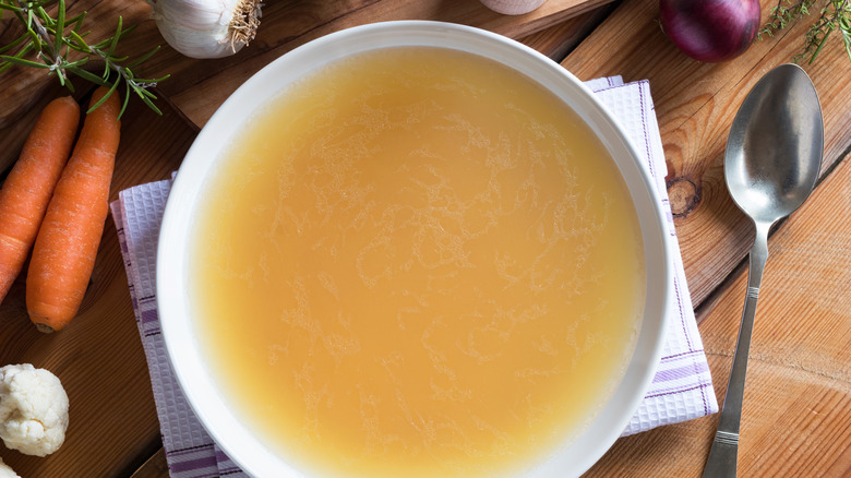 A bowl of chicken stock with spoon and carrots nearby