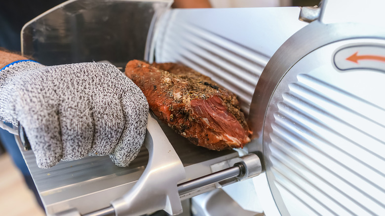 A worker wears a protective glove while cutting meat with a slicer machine.