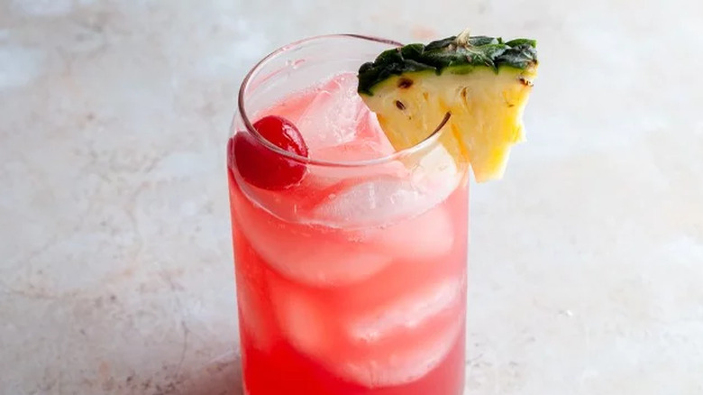 Tall glass with red cocktail with ice, cherry, and pineapple piece.