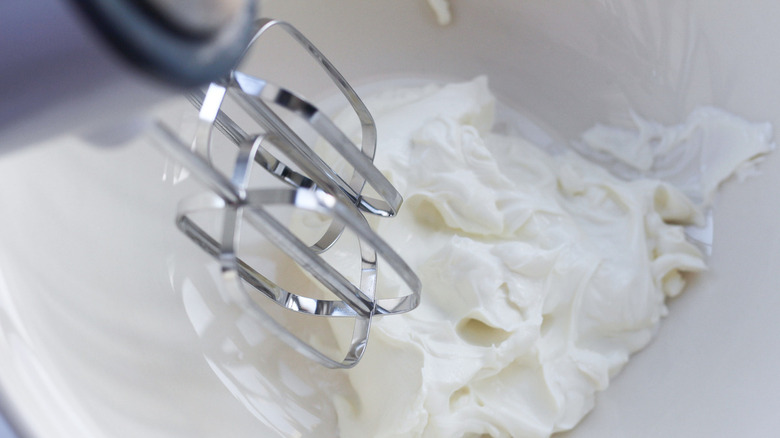 An electric whisk's beaters above a bowl of cream cheese