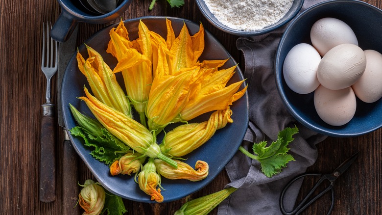 squash blossoms line a blue bowl on a wood surface surrounded by other ingredients