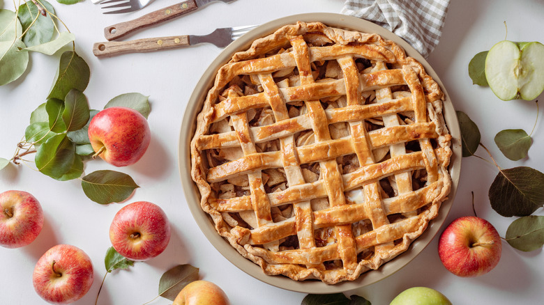 An apple pie surrounded by apples on a table