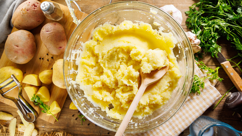 Mashed potatoes in bowl with whole potatoes and herbs on table