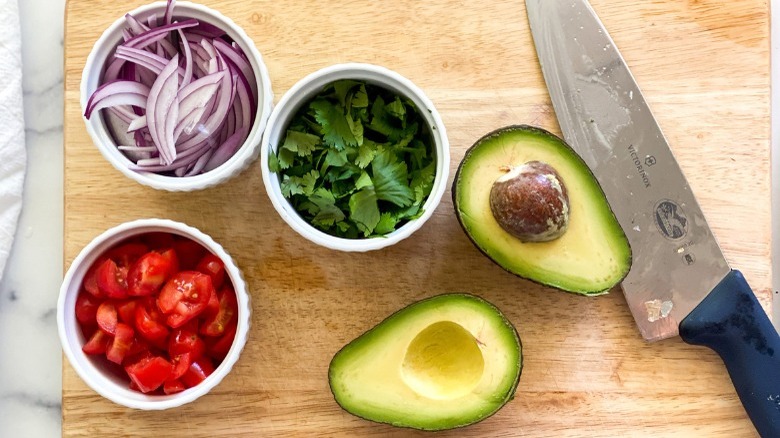 bowls of sliced red onion, cilantro leaves, and diced tomato; halved avocado; knife on cutting board