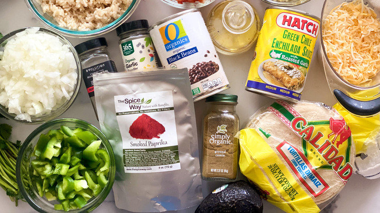 A can of black beans surrounded by rice, onions, red and green peppers, oil, corn, cheese, tortillas, cumin, enchilada sauce, garlic, salt and paprika