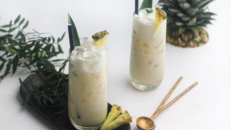 Two light yellow cocktails with pineapple and whipped cream on a white background