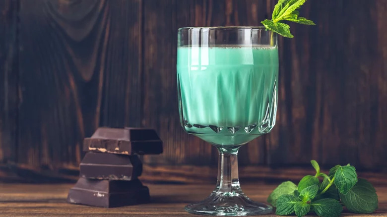 A creamy green drink in a goblet, with chocolate ad mint leaves next to it