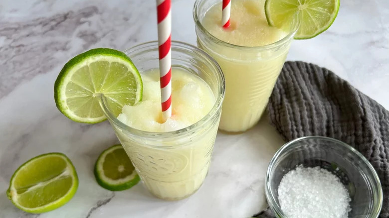 Two frozen margaritas in jars, next to sliced limes and a bowl of salt