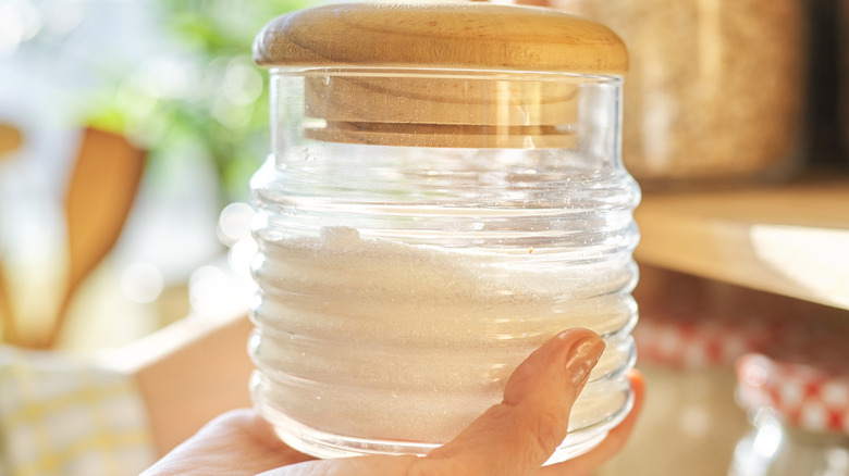 hand holding jar of white sugar with pantry shelves in background