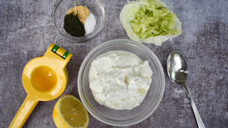 A hand held lemon juicer next to a zested lemon, a bowl of spices, a bowl of Greek yogurt, some grated cucumbers, and a spoon