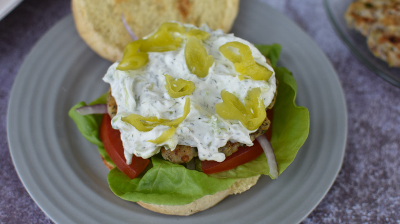 An open chicken burger with lettuce, tomato, tzatziki, red onions, and banana peppers on a blue plate