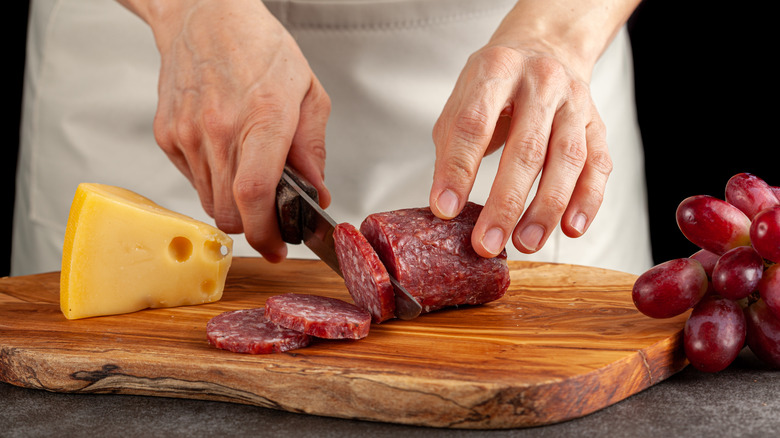 jarlsberg on a cutting board next to salami being sliced