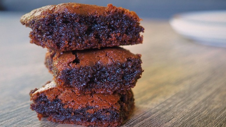 Three gooey brownies stacked on a wood surface.