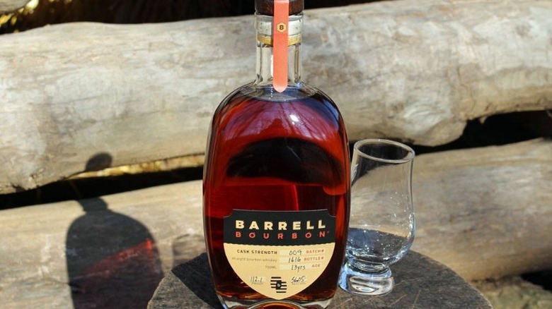 bottle of Barrell Bourbon and whiskey glass resting on logs 