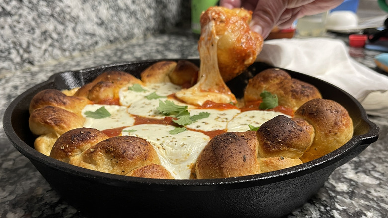 hand holding a bread knot above a black frying pan filled with cheese and tomato sauce