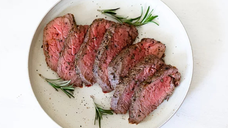 Slices of rare beef tenderloin on plate with rosemary sprigs.