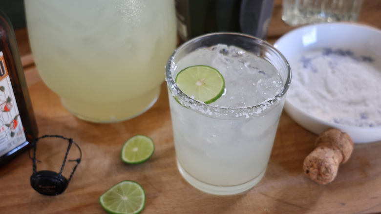 champagne margarita in rocks glass with salted rim surrounded by pitchers, bottles, and a champagne cork