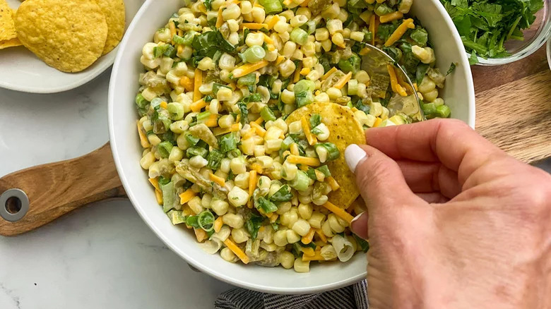 hand dipping tortilla chip into bowl of creamy corn dip