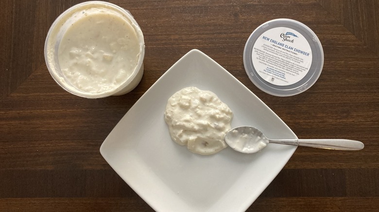 Some Blount's New England Clam Chowder is on a plate with a spoon and next to a soup container.