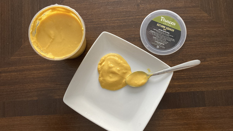 Some Panera Autumn Squash soup rests on a plate with a spoon next to the soup container.container