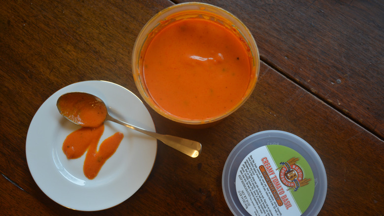 Some creamy tomato basil soup is on a plate with a spoon next to a soup container.