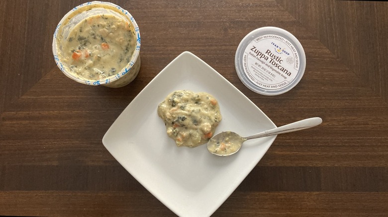 Some Ivars Rustic Zuppa Toscana soup is on a plate with a spoon and sits next to a soup container.