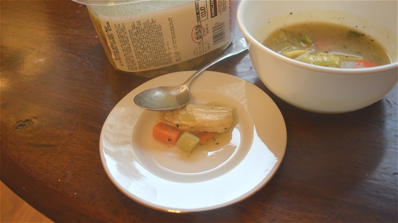 Some Rotisserie Chicken Soup sits on a plate with a spoon next to a bowl and soup container.