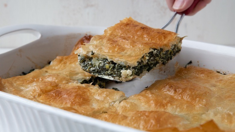person serving square of spanakopita from casserole dish