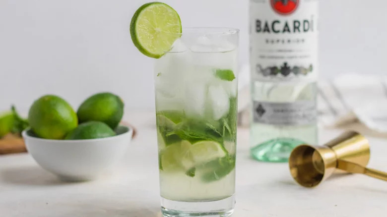 mojito in glass with cucumber garnish and bottle of bacardi and limes in background