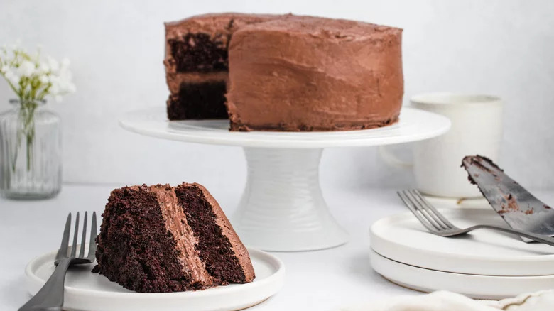 A slice of chocolate cake with a chocolate cake on a stand in the background