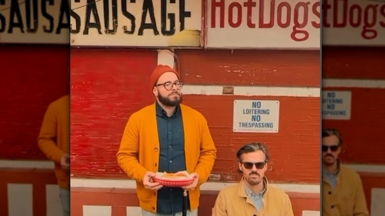 Two men with a hot dog basket against a red wall.