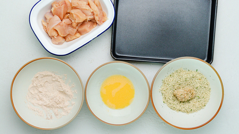 three bowls of flour mixture, seasonings, and egg mixture with diced chicken bites near a tray