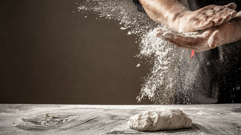 disc of dough on a floured surface with a man's hands throwing flour