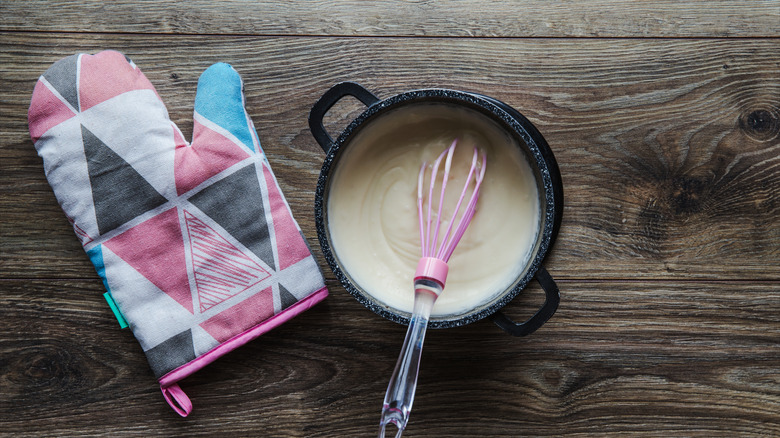 White béchamel sauce in a small black saucepan on a wooden table with a pink whisk for stirring the sauce and a pink pot holder with an abstract pattern