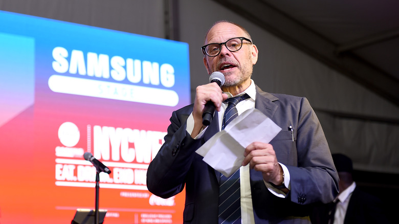 Alton Brown speaking at a microphone while wearing suit and tie