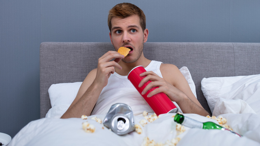 Man in bed with beer cans, bottles, chips, and popcorn