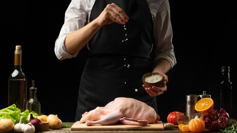 Cook in black apron, set against black background, sprinkling salt on raw whole duck on a cutting board, surrounded by ingredients like garlic cloves, oranges and oils.