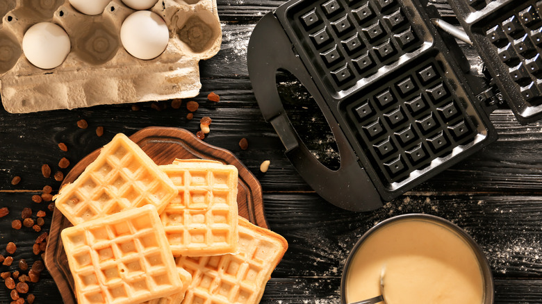 square waffles, waffle maker, batter, and carton of eggs on wooden table