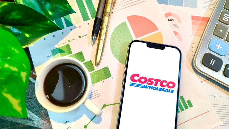 Costco app is visible on a phone lying on a table with coffee, pens, and papers.