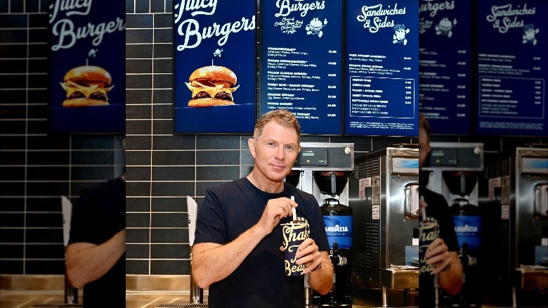 Bobby Flay holding milkshake in front of bobby's burgers menu