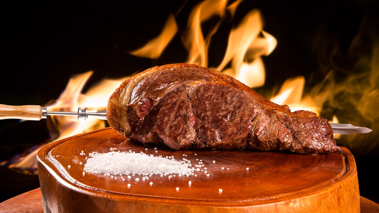 slab of skewered meat on wooden plant with salt in front of fire