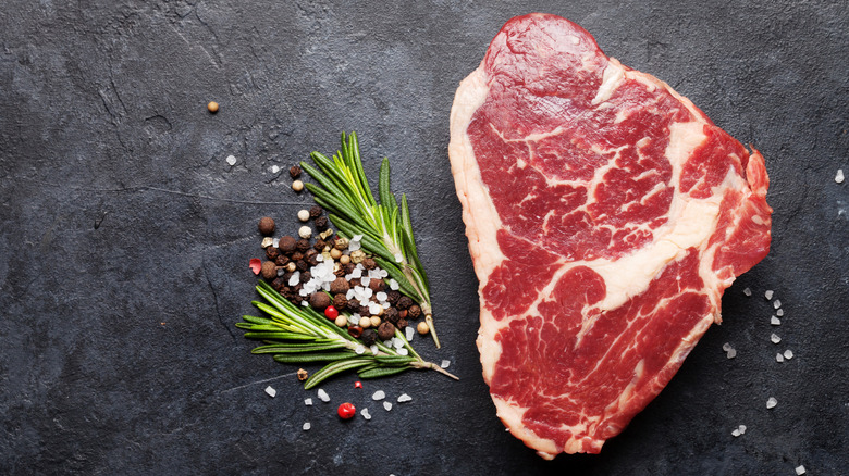 raw ribeye steak next to salt, peppercorns and rosemary