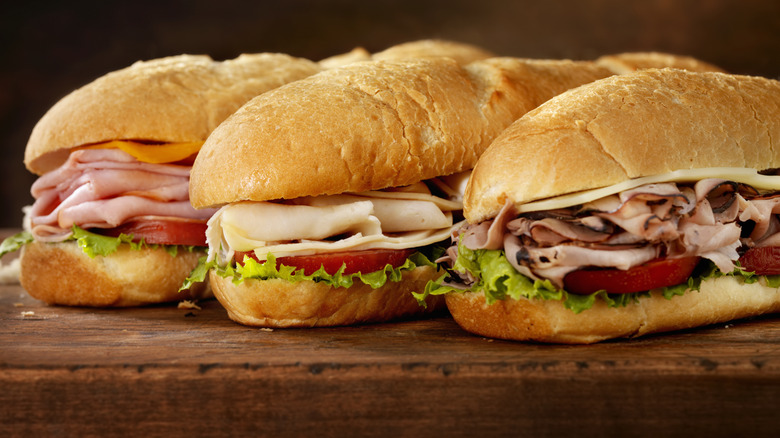 Three sub sandwiches aligned on a wooden cutting board