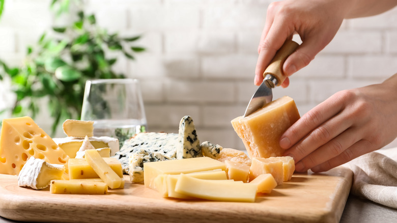 Hands cutting a parmesan cheese next to a board with more cheeses