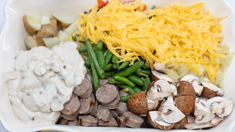 Overhead shot of a white baking dish filled with cream of mushroom soup, potatoes, cheese, green beans, bratwurst, mushrooms, and onions