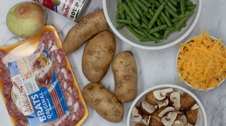 A package of bratwurst next to an onion, a cane of cream of mushroom soup, four potatoes, a bowl of green beans, a bowl of shredded cheese, and a bowl of mushrooms