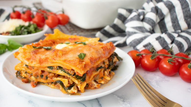 lasagna on a white plate surrounded by tomatoes and black-striped napkins