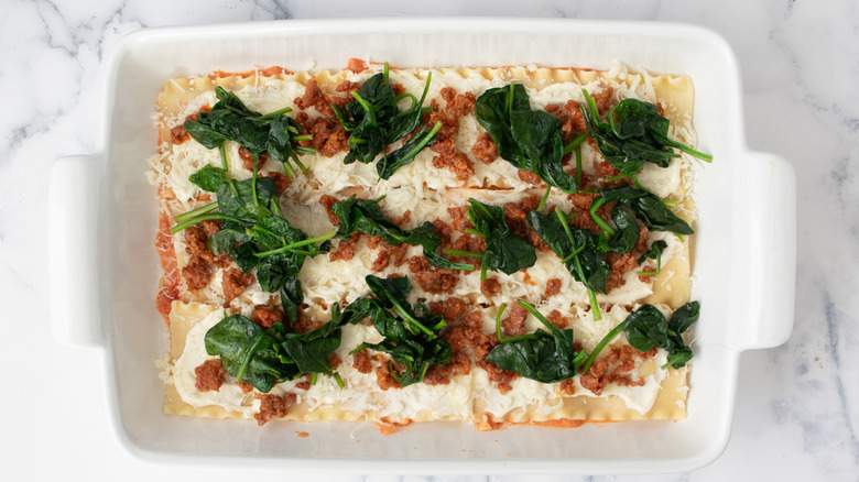 lasagna noodles covered with white paste, ground meat, and spinach in a white baking pan