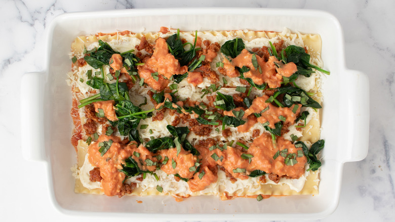 lasagna noodles covered with white paste, ground meat, pinkish-orange sauce, and spinach in a white baking pan