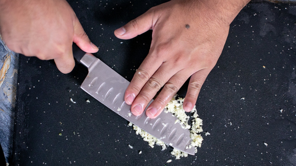 hands crushing minced garlic with a knife on a black cutting board